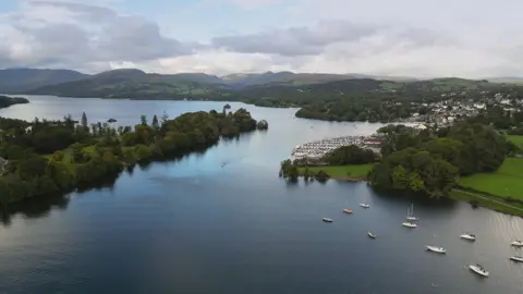 An aerial image of the lake at Windermere, with boats sailing and others moored at a marina. The town, with its houses and buildings are also visible in the distance. Further away there are the fells and green spaces of the Lake District.