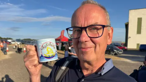 Phillip Norton/BBC. Ian Paterson aparece en la foto con una taza de Hykeham Sailability. Tiene el pelo corto y gris, lleva gafas y viste una camisa polo azul.