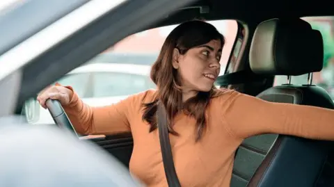 A woman sits in her car and looks backward to reverse it. She is wearing a seatbelt and has one hand on the wheel, with her arm on the passenger seat. Another car can be seen through the window.