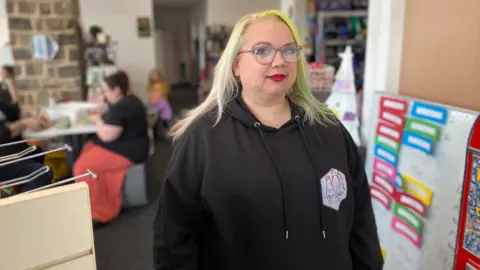 A woman with long blonde hair, some of dyed green, wearing glasses and red lipstick and a black hoodie, smiles at the camera. In the background are people playing table-top games.