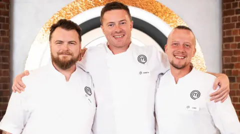 BBC/Shine TV The three finalists from left to right, Mark O'Brien, Gareth Baty and Luke Emmess. All three chefs are wearing white chefs' coats and black trousers. O'Brien has short dark hair and a full beard. Baty, who has his arms round the shoulders of the other two, is taller and has similar short dark hair with light stubble. Emmess has very short dark hair on the top of his head and is shaved at the sides. He has a short goatee beard. They are on the set of MasterChef, with a brick wall and the programme's logos behind them.