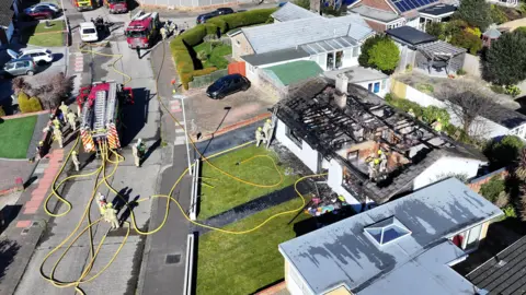 A white bungalow in a residential street is seen from above. It's roof is completely burnt. 