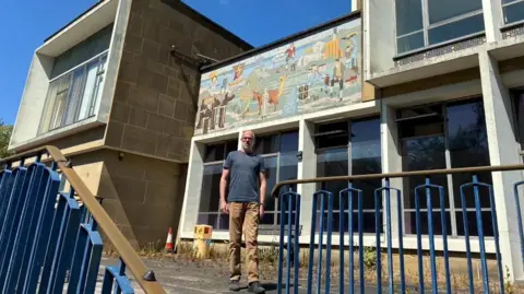Supplied Bennett Zon, in light brown trousers and a grey t-shirt, standing in front of the mosaic mural. The artwork sits above tall windows outside Durham County Hall. Weeds are growing from the foundation of the building, which is being emptied ahead of demolition. 