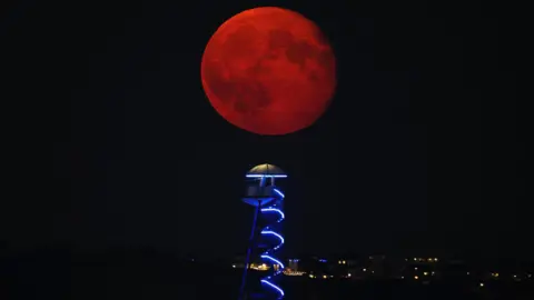 A red-looking moon can be seen above a helter skelter lit up in the dark on Bournemouth seafront. Lights from buildings can be seen in the dark background.