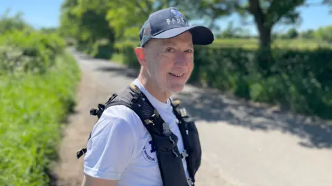 Peter smiling at the camera wearing a dark baseball cap and white T-shirt underneath a black running vest with water bottles inside its pockets. He is stood in a layby on a country lane, with blurred hedges and trees in the background.