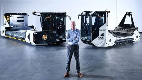 A man standing an a dark warehouse that has dim lighting. He is standing with arms crossed in front of two self-driving vehicles that are used to move airport baggage. He is wearing suit trousers and a blue shirt and has short grey hair. He is smiling. 
