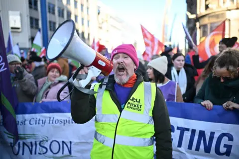 A union steward in a high-viz jacket and pink hat shouts while holding a megaphone to his mouth. Behind him a crowd with red and purple flags can be seen, a banner is being held up with the blue Unison logo on a white background