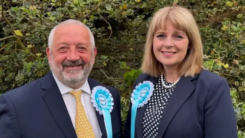 Reform UK A man and a woman are wearing light blue and white rosettes featuring the Reform UK logo. The man, on the left with grey hair and a beard, and the woman, with blonde/brown hair, are both wearing blue jackets.