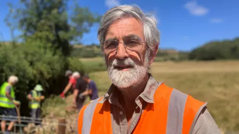 An image of Mick Fletcher, Chair of the Strawberry Line. He is has grey hair and a grey beard and is wearing silver rimmed glasses. He has a beige shirt on with an orange high-vis vest over it. He is standing in a field with people wearing high vis vests in the background. 