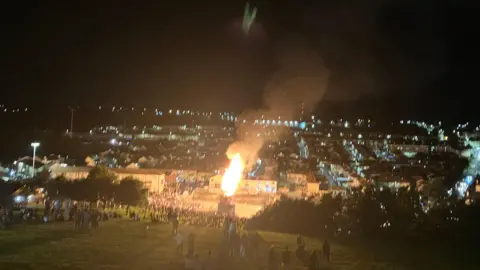 A large fire is visible from a high point and the lights of the city are behind it