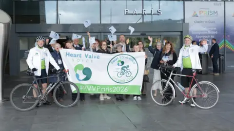 Royal Sussex County Hospital Two cyclists standing for a photo outside a building with people cheering them on stood between them