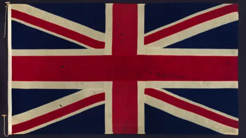 A Union Flag in a frame. It has a number of small holes in the fabric.