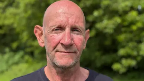 Lee is stood looking at the camera, wearing a dark tshirt. He is shaven headed with short, stubbled facial hair. He is outdoors on a sunny day, with a green hedge blurred in the background.