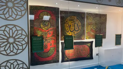 Tabassam Zabier A glass cabinet, lit, features four colourful prayer mats 