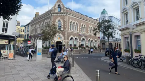 People walk around the Broad Street area of St Helier on a sunny but cloudy day. Three people are sat on metal benches along the road. Several bicycles are locked onto bike rakes on both sides of the street.