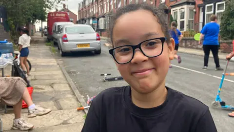 A seven year old boy wearing a black t shirt and black glasses is smiling at the camera in the centre of frame. Behind him are children playing on scooters and with tennis bats and balls in a residential street, while parents sit on the pavement. 