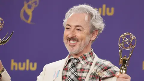EPA Alan Cumming wearing a cream jacket with a tartan shirt and tie. He is smiling at the camera holding two trophies.