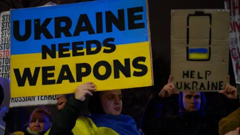 Getty Images Demonstrators hold a signs at a protest outside the US embassy in Warsaw on 3 March, 2025. A man wearing a Ukrainian flag, hold a sign painted with the flag as the background and the words: Ukraine needs weapons, in black lettering. To the left of the man is another, holding a sigh with a nearly empty battery painted on it, filled slightly with the flag and the words Help Ukraine.