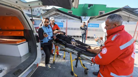 Reuters A patient is brought to an ambulance as he gets ready to leave Gaza for treatment abroad through the Rafah border crossing between Gaza and Egypt after it was opened by Israel on Monday for a limited number of people, in Khan Younis in the southern Gaza Strip, February 5, 2026. Two medical workers in orange are moving him to the vehicle. He's lying on a stretcher, covering his face with his left hand. 