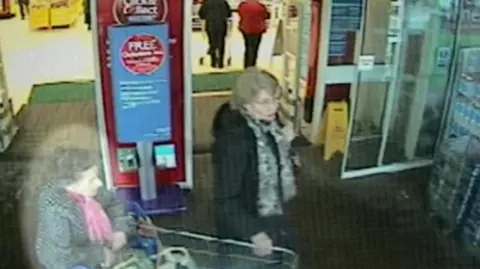 Cambridgeshire Police Blurry CCTV image showed Una Crown on the left wheeling a shopping trolley. She is wearing a coat and a pink scarf. Another woman in a dark coat and grey scarf is helping with the trolley