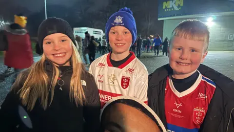 Three children, a girl and two boys, smile at the camera. They are wearing Hull KR merchandise and are holding a cardboard cut out of a player.