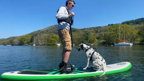 Jonathan London Jonathan London stands on top of a paddleboard which has a dalmatian dog also sitting on it. The paddleboard is bright green and is looks like a hot day on Windermere.