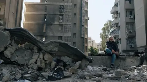 A man sits on a concrete, surrounded by rubble.