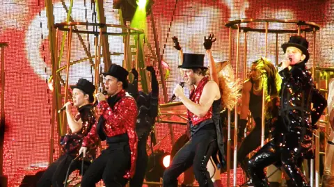 Getty Images Take That are pictured performing on stage. The four members of the banned are wearing glittery red tops and black top hats. They are all singing into microphones in front of a large electronic screen. 
