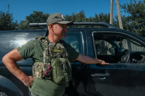 Andrey Ohinok, known as a Jackie Chan Volunteer, gazes off to the right, standing beside a black car, holding onto the window frame. He has camouflage gear on, a camo rucksack, brown binoculars and a khaki skip hat.