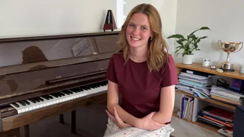 Freya Terris is a young woman with shoulder length light brown hair, which is hanging in loose waves around her face. She is smilling at the camera. She is wearing a burgundy t-shirt and is sitting on a piano stool with her arms crossed. She is sitting in front of a glossy brown piano, and there is a metronome, a plant and a large trophy seen sat around her. 