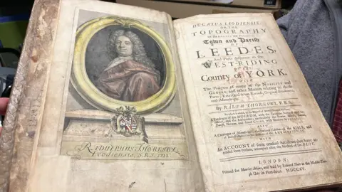 Leeds City Council The image shows a very old, worn book being held open by a person. The book appears to be an early 18th-Century historical work, with aged yellowed pages and a damaged binding.