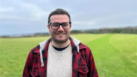 BBC Man in black rimmed glasses and checked shirt smiles at camera while standing in a green field