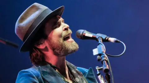 Gaz Coombes of Supergrass is wearing a fedora and is singing into a microphone.