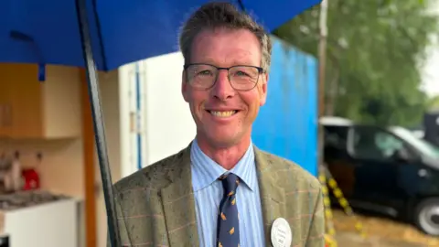 A man wearing a green blazer and blue shirt, he has dark hair and glasses