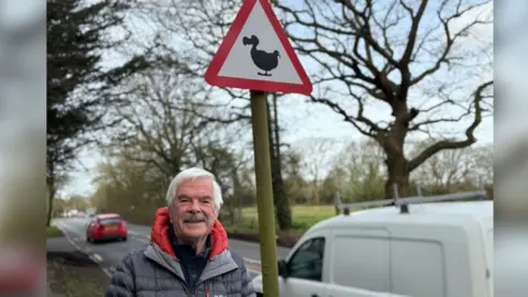 A man smiling straight at the camera and standing next to a road sign with a dodo on it.