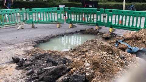 A hole in a road filled with water surround by rubble and green fencing