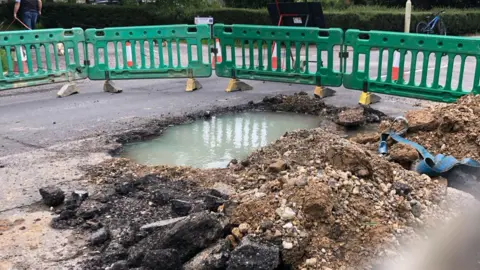 A hole in a road filled with water surround by rubble and green fencing