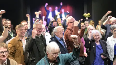 Paul Moseley/BBC A group of people raising their hands in celebration. In the background three figures are standing on a stage with their hands raised.
