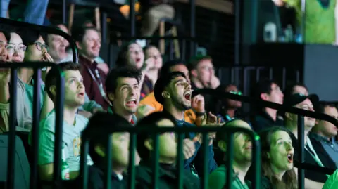 MEWC Audience members sat in a tiered seating area looking shocked, apprehensive and shouting at results for the MEWC finals, off screen. They are bathed in a green light.