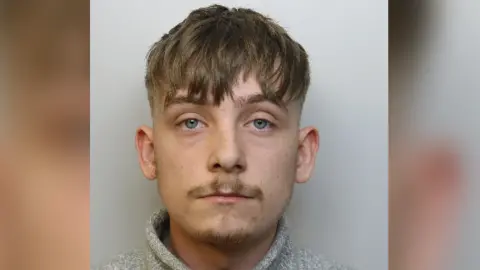 A mugshot of Taylor Stacey. He is wearing a grey jumper and has short mousey brown hair with blue eyes and a solemn look on his face.
