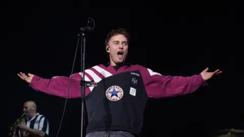 PA Media Sam Fender on stage. He is wearing a Newcastle United top which contains the club logo as well as the Newcastle Brown Ale star. A saxophone player in the background is wearing a Newcastle United shirt.