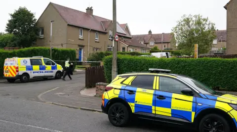PA Media Police at crime scene in Edinburgh. Two police cars are packed at the end of a street of small, brown-harled houses. There is police tape over the entrance to the street.