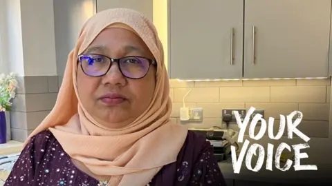 A woman in a peach hijab and glasses stands in a modern grey-hued kitchen. 