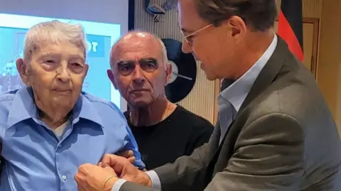 German Embassy in Israel Michael Smuss, with white hair and wearing a blue shirt and tan trousers, stands up on a walker as a man with dark hair in a suit pins a medal to his chest.