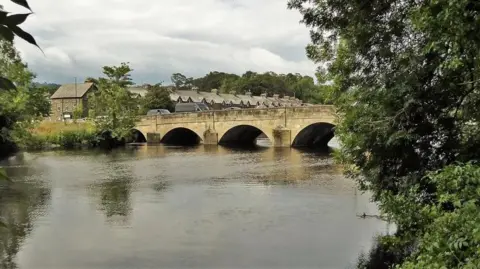 Kevin Waterhouse/Geograph A bridge spanning a river, pictured from across the water. There are some trees to the side. 