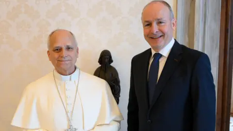 Vatican Media Pope Leo standing, wearing all white, standing beside Taoiseach Micheál Martin, wearing a navy suit and white shirt. 