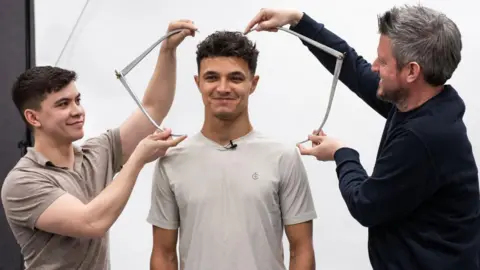 Image of two men, standing on either side of a third man. Both men are holding calipers and measuring the third man's head.