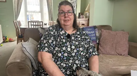 Oliver Whitfield-Miocic/BBC Nikki Paine pictured at her home in Ashford, Kent. She is sat on a beige sofa and has a dog at her feet. She is wearing a floral dress and has long brown hair and glasses.