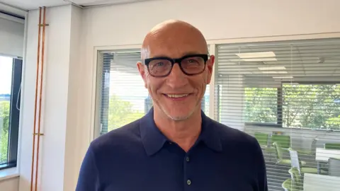 A photo of a man who is bald and has stubble. He is wearing glasses and a blue polo shirt. He is stood in an office with a window and chairs in the background.