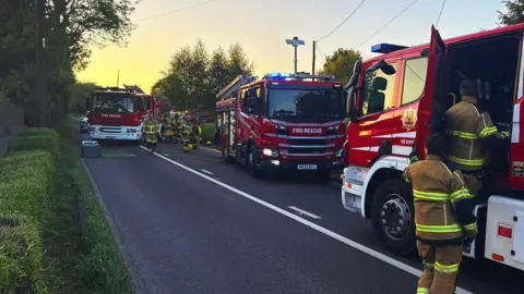 Shropshire Fire and Rescue Service Fire engines are parked along a nearby road, blocking the carriageway. Firefighters wearing uniform are working around them.